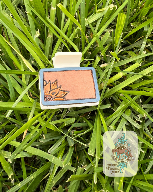 Cream base hair clip with a tooled and painted sunflower and a blue border. Photographed on grass with logo in bottom right corner of photo.