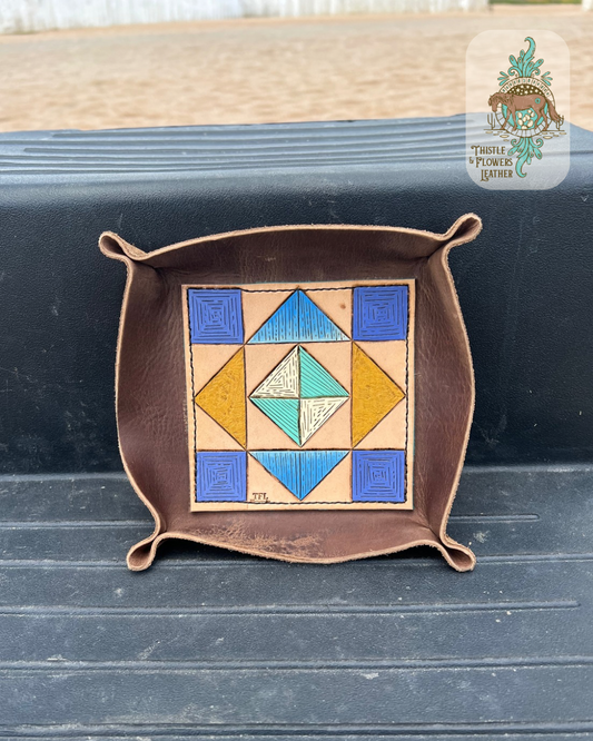 Valet tray with a tooled and painted leather base. Base is tooled and painted to appear like a barn quilt or quilt square. Painted colors are dark blue, light blue, yellow, white, & turquoise. Tray is on a mounting block step. Logo is in top right corner of photo. 