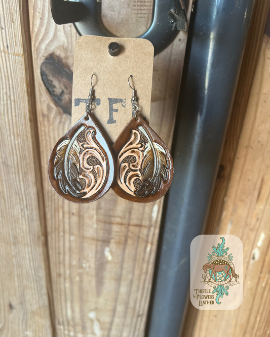 Tooled leather teardrop shaped earrings with traditional Western tooling and a feather on each earring. Feathers are painted in various shades of cream, browns, and black. Earrings are hand tooled and painted. Earrings are hanging on stall front. Logo is in bottom right corner of photo.