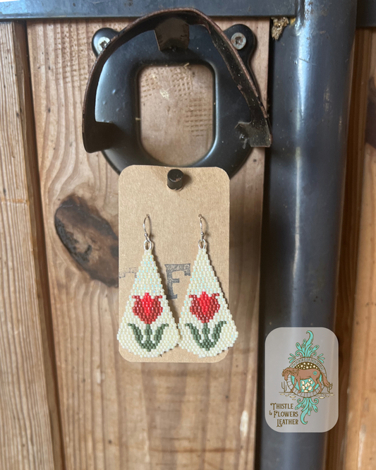 Hand beaded earrings with a vintage flower design. Bead colors are red, a lighter red, cream, and green. Earrings are a longer teardrop shape. Earrings are hanging in stall front. Logo is in bottom right of photo.