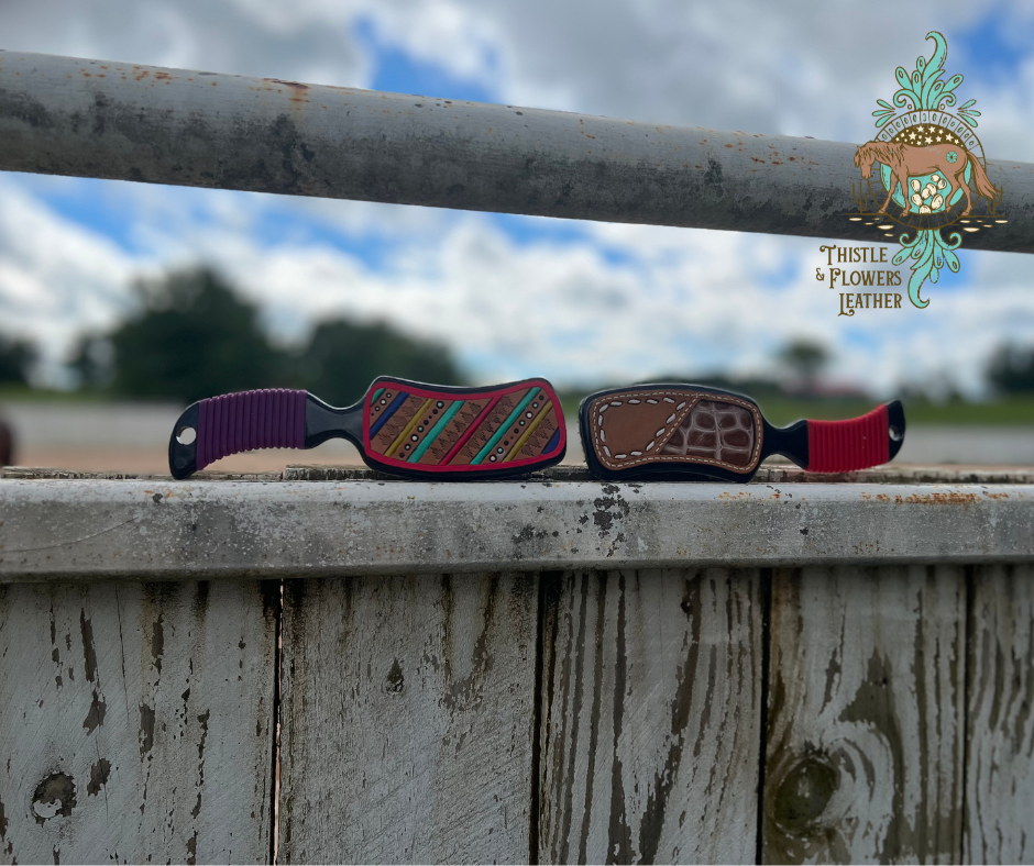 2 brushes on an arena fence post. The left one has a purple grip and a tooled leather patch with a hot pink outline and a western design with various colors like yellow, dark blue, turquoise, and hot pink. The one on the right has a red grip, and a leather patch with a faux buckstitch pattern and a natural colored croc inlay..