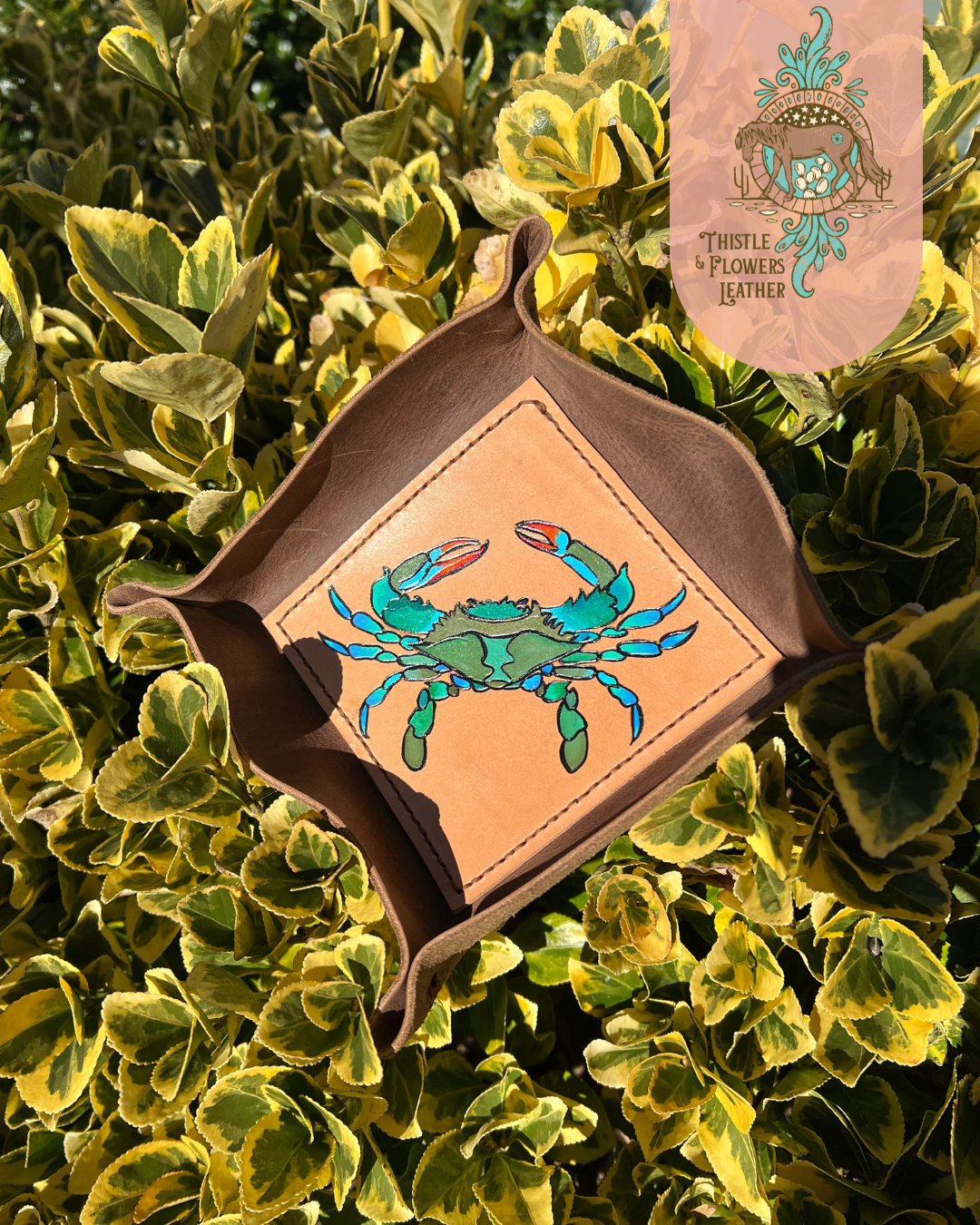  Leather Valet tray with painted blue crab on bottom of tray. Tray is propped up in a green and yellow bush, with strong golden hour sunlight highlighting colors.  Photo is watermarked with the Thistle & Flowers Leather logo.