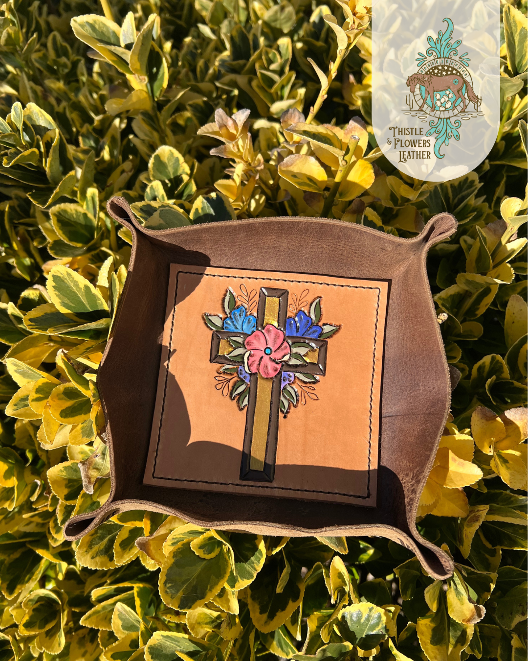  Leather Valet tray with tooled and painted cross with flowers around it on bottom of tray. Tray is propped up in a green and yellow bush, with strong golden hour sunlight highlighting colors and metallic paint in cross.  Photo is watermarked with the Thistle & Flowers Leather logo.