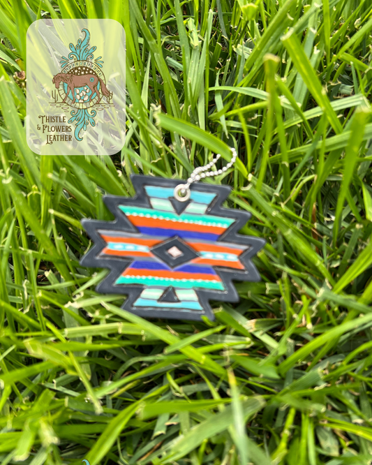 Southwestern shape leather keychain that has been tooled with a border and diamond in the center and painted with stripes. Paint colors are different shades of rust, turquoise, blue and white. Keychain is on grass. Logo is in top left of photo.
