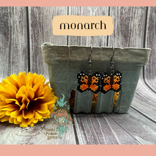 Small beaded earrings in the shape of a butterfly. Bead colors are light orange, orange, black, and white. Looks like a pixelated monarch butterfly. Black shiny earring hooks and jump rings. Earrings are hanging on a vintage green paper produce container, with a yellow fake marigold flower next to it. Backdrop is gray wood. “Monarch” is in a text box with peach background to identify style, and the Thistle & Flowers Leather logo is on the flower and produce container.