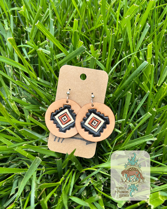 Round tooled and painted leather earrings with a silver stud. Earrings are tooled with a southwestern design, and painted with black, rust and cream colors. Photographed on grass with logo in bottom right corner of photo.