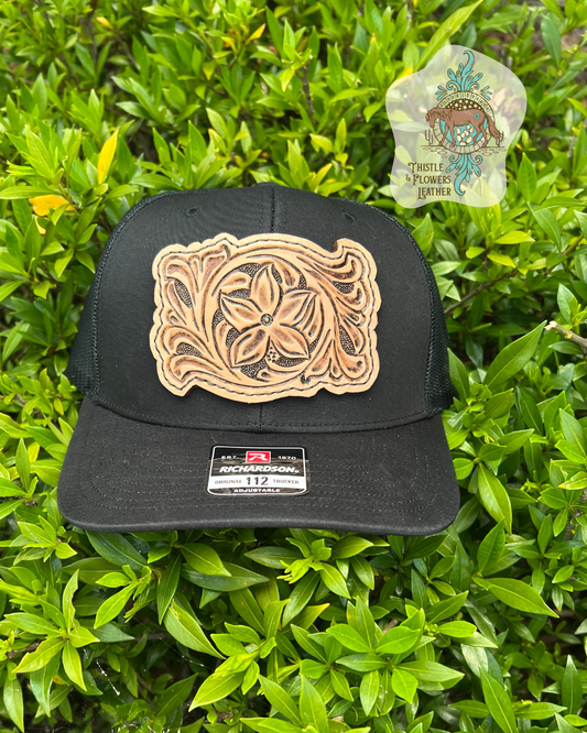 Black richardson 112 hat with a tooled leather patch stitched onto hat with dark brown thread.  Leather Patch has a flower and traditional western vines tooled on it. Business logo is in upper right corner of picture, not on hat. Hat is resting on bright green bush.