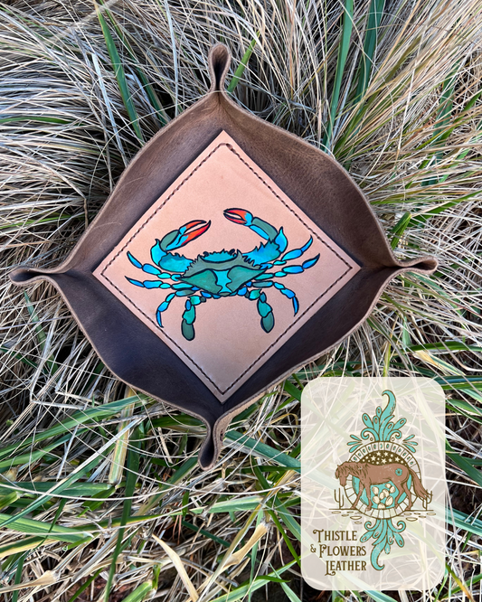 Leather Valet tray with painted blue crab on bottom of tray. Tray is laying the taller, clean grass. Photo is watermarked with the Thistle & Flowers Leather logo. 