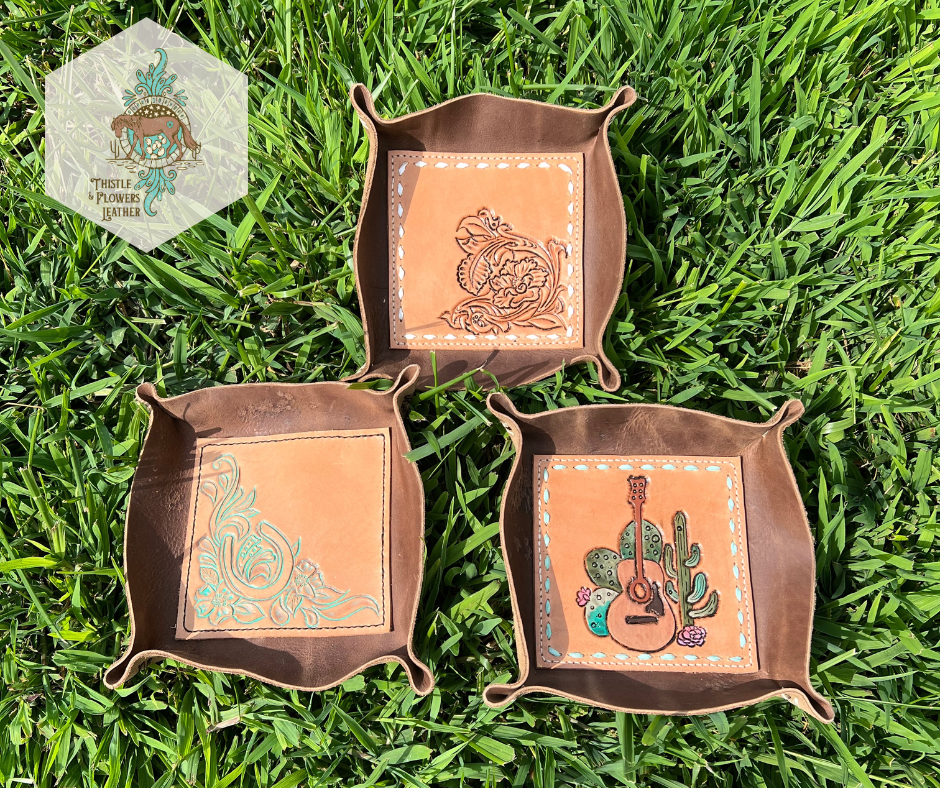 3 Leather valet trays, all with chocolate colored leather exterior and a tooled veg tan center. The top tray is floral and scroll tooling with white buckstitch. The bottom left is a tooled horseshoe with flowers and scrollwork. The bottom right is a tooled and painted acoustic guitar with cacti around it, with mint suede buckstitch.