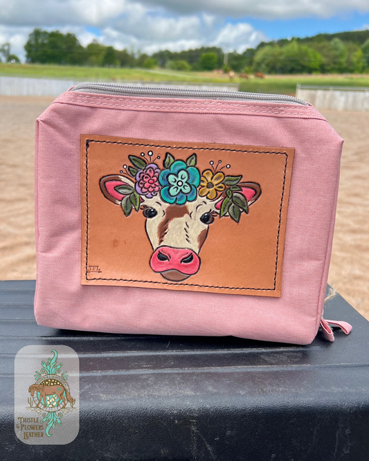 Pink makeup bag with a hand tooled and painted leather patch featuring a cow wearing a flower crown. Flowers are shades of yellow, blue, and pink/purple, with leaves in shades of green. Cow has pink nose and inner ear and a sorrel and cream piebalding pattern. Makeup bag is sitting on a mounting block in an arena. Logo is on the bottom left of photo.