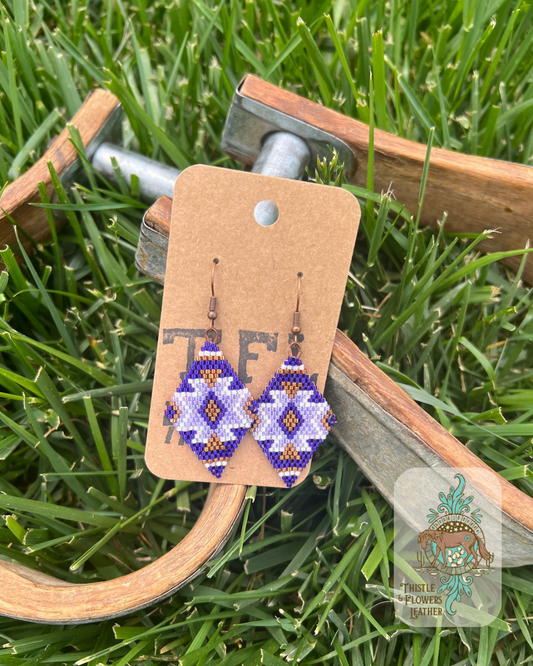 Hand beaded earrings in a diamond shape with a southwestern design. Beads are dark purple, light purple, metallic copper, and white. Photographed on wooden and metal stirrups on grass. Logo is in bottom right corner of photo.