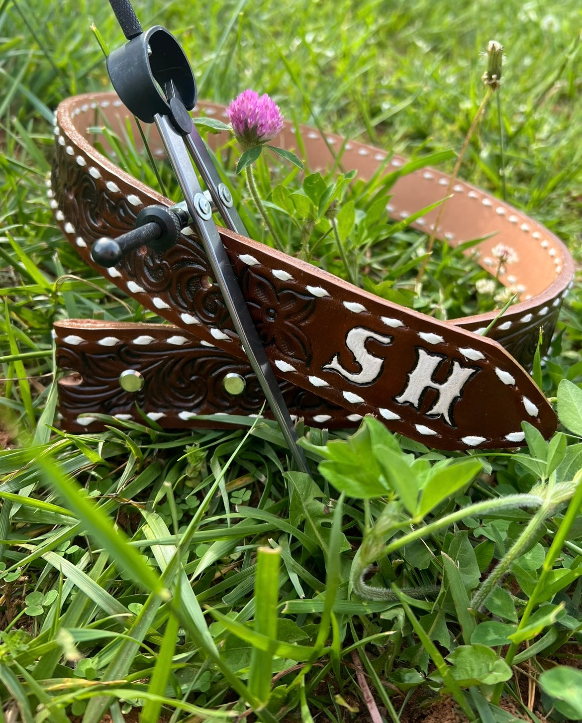 Custom tooled in a traditional western floral style belt. Belt is dyed a dark brown with white buckstitch. Belt has 2 western initials on the end, “SH”. Belt is being held in a circular shape by a leather tool. Belt is one grass with a purple clover flower