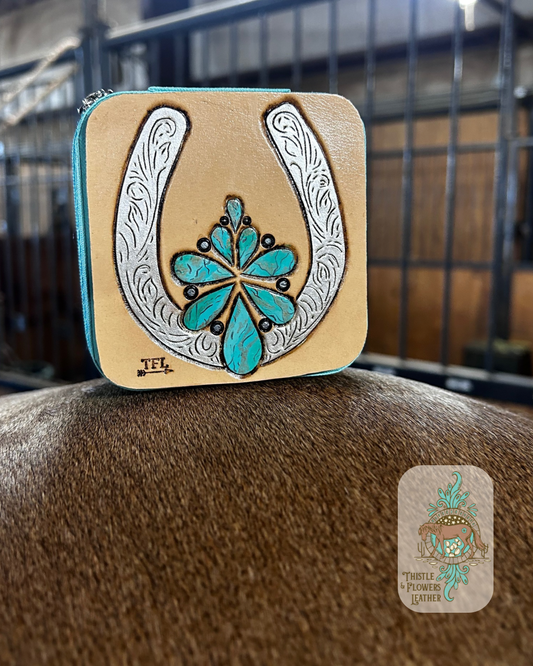Mini travel jewelry box with a hand tooled and painted leather patch. Patch features a tooled and painted faux turquoise squash blossom and a tooled and painted silver horseshoe with detail carving. Jewelry box is sitting on a horse in a barn. Logo is on bottom right of photo.