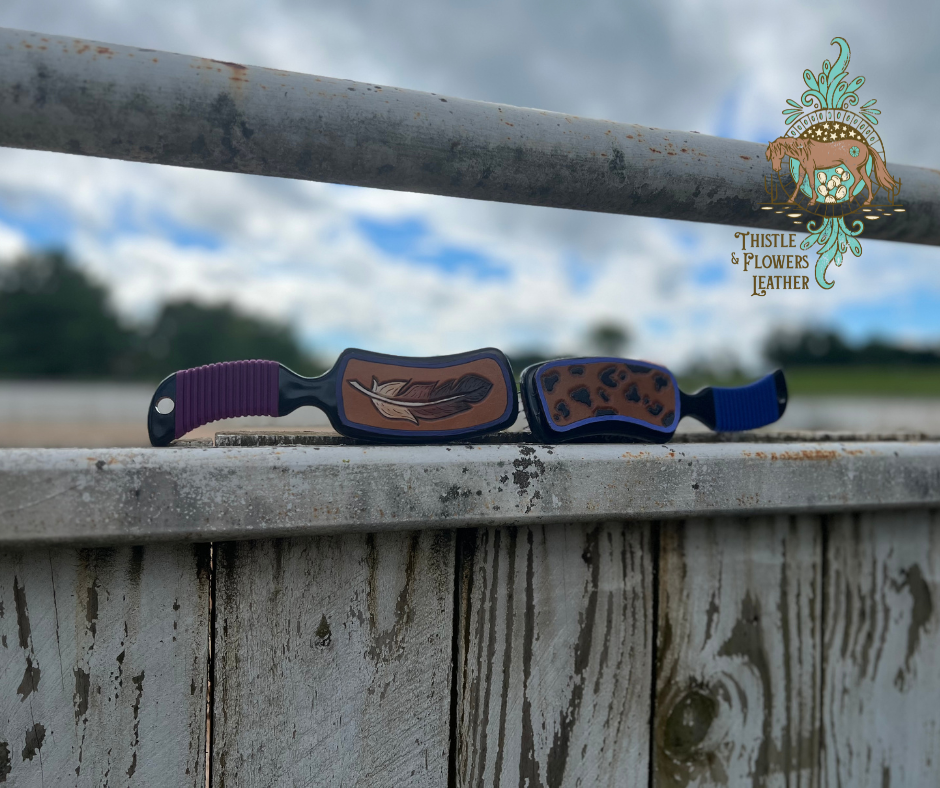 2 brushes on an arena fence. the one on the left has a purple grip and tooled leather patch with a dark purple border and an intricate feather tooled and painted with an ombre effect, with white at the base of the feather, and black at the tip of the feather. The brush on the right has a blue grip, with a periwinkle border and a cheetah print tooled and painted.