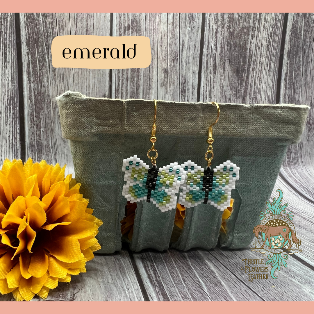 Small beaded earrings in the shape of a butterfly. Bead colors are light green, teal, turquoise, black, and white. Looks like a pixelated butterfly. Gold colored earring hooks and jump rings. Earrings are hanging on a vintage green paper produce container, with a yellow fake marigold flower next to it. Backdrop is gray wood. “Emerald” is in a text box with peach background to identify style, and the Thistle & Flowers Leather logo is on the flower and produce container.