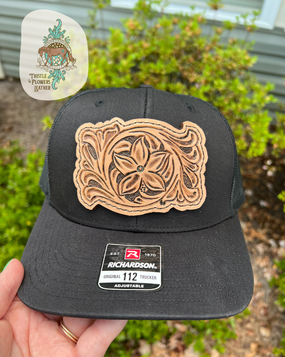 Black richardson 112 hat with a tooled leather patch stitched onto hat with dark brown thread. Leather Patch has a flower and traditional western vines tooled on it. Business logo is in upper left corner of picture, not on hat. Hat is being held in front of bushes.