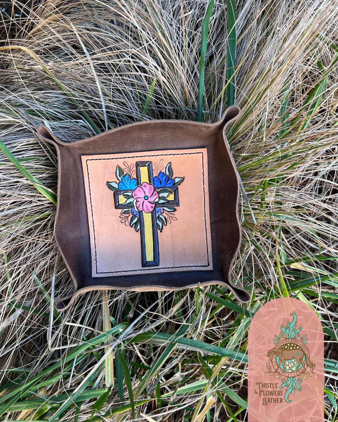  Leather Valet tray with tooled and painted cross with pink, light blue, and dark blue flowers around it on bottom of tray. Tray is laying in tall clean grass. Photo is watermarked with the Thistle & Flowers Leather logo.