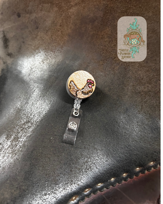 Badge reel with a tooled and painted leather patch. Leather patch has a chicken tooled and painted on it. Chicken colors are various shades of brown, cream, yellow, and red. Reel is sitting on a saddle seat. Logo is in top right corner.