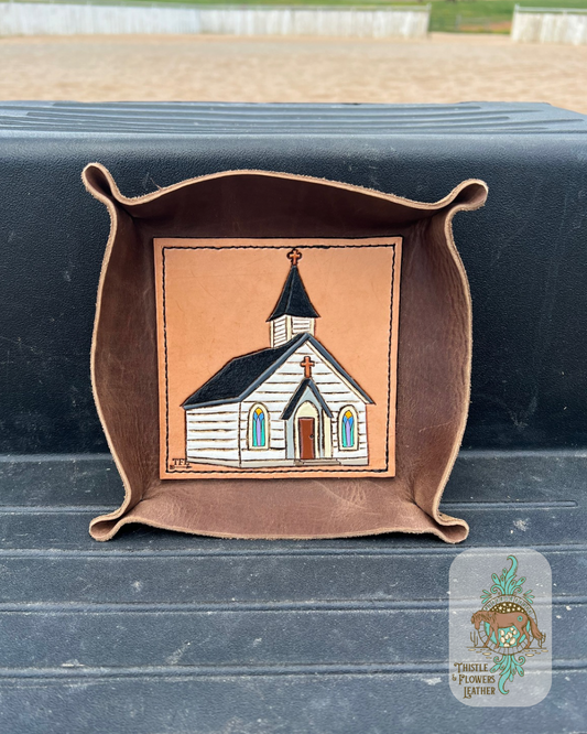 Brown leather valet tray with a hand tooled and painted leather base. Base has a white chapel with stained glass windows, brown door, and black roof tooled and painted on it. Tray is sitting on mounting block. Logo is in bottom right of photo. 