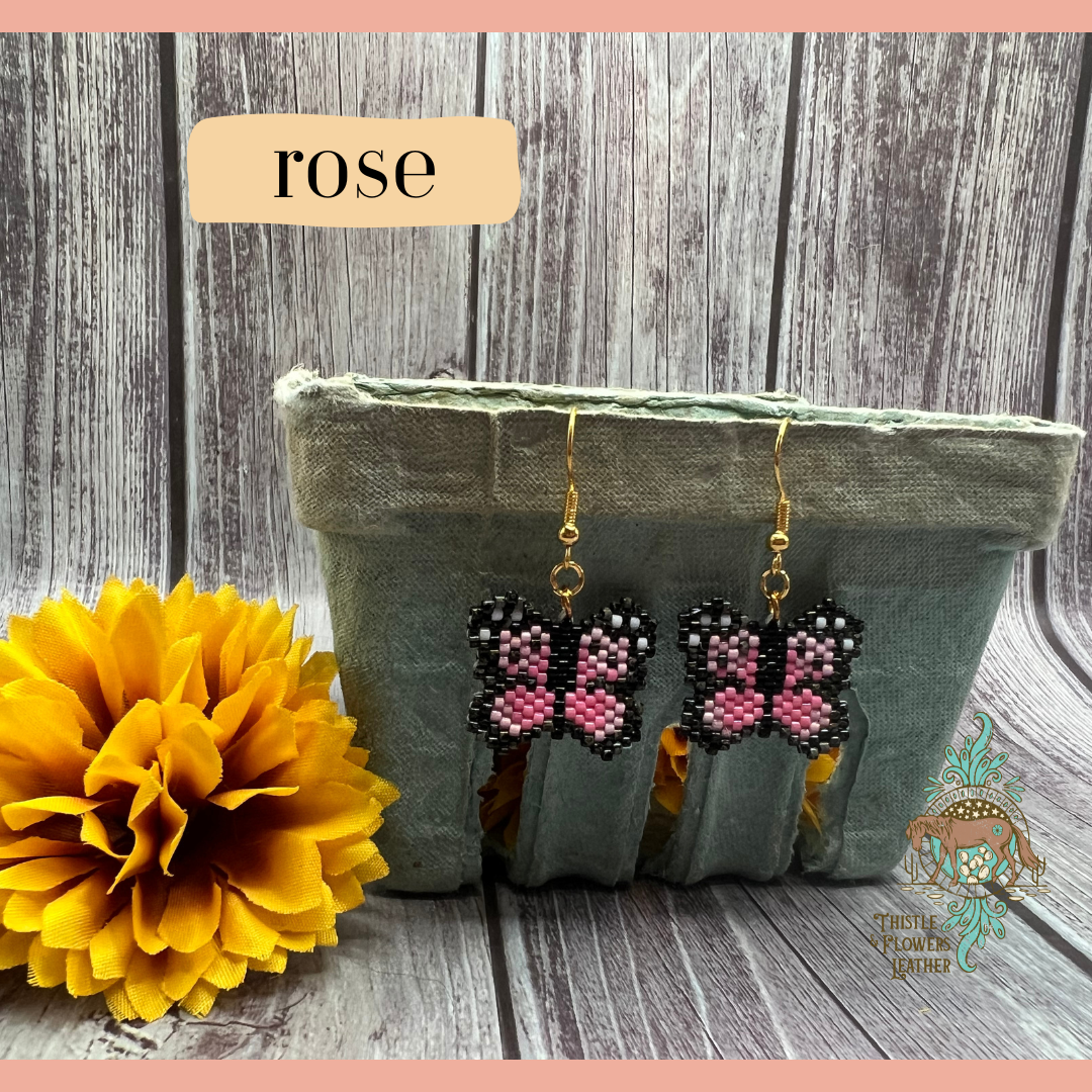 Small beaded earrings in the shape of a butterfly. Bead colors are light pink, pink, black, and white. Looks like a pixelated butterfly. Gold colored earring hooks and jump rings. Earrings are hanging on a vintage green paper produce container, with a yellow fake marigold flower next to it. Backdrop is gray wood. “Rose” is in a text box with peach background to identify style, and the Thistle & Flowers Leather logo is on the flower and produce container.