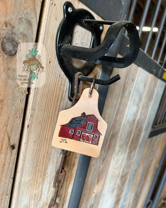 Cowtag shaped tooled leather keychain with a tooled and painted red barn on on it. Keychain is hanging on a stall door. Logo is on the top right of the photo.
