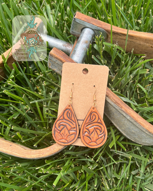 Teardrop shaped tooled leather earrings with a flower and traditional western tooling. Photographed on metal and wooden stirrups on grass, with logo on top left corner of photo.