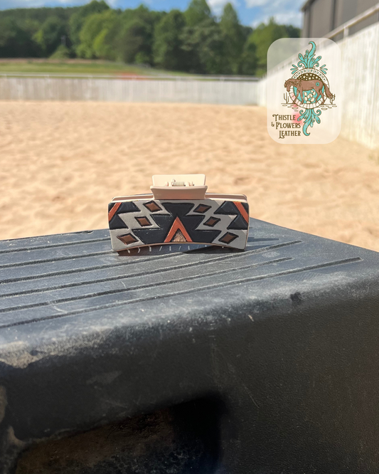 Large hair clip with cream colored base and a hand tooled and painted patch. Patch has a southwest design tooled and painted. Colors are gray, black, rust, and brown. Clip is on a mounting block and in an arena. Logo is on the top right of photo.
