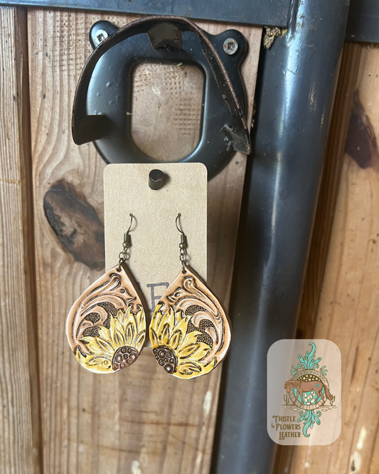 Hand tooled and painted teardrop leather earrings. Earrings have traditional western carving and sunflowers peeking in from a bottom edge. Sunflowers are painted 2 shades of yellow with a brown center. Earrings are hanging on stall front. Logo is on bottom right of photo. 