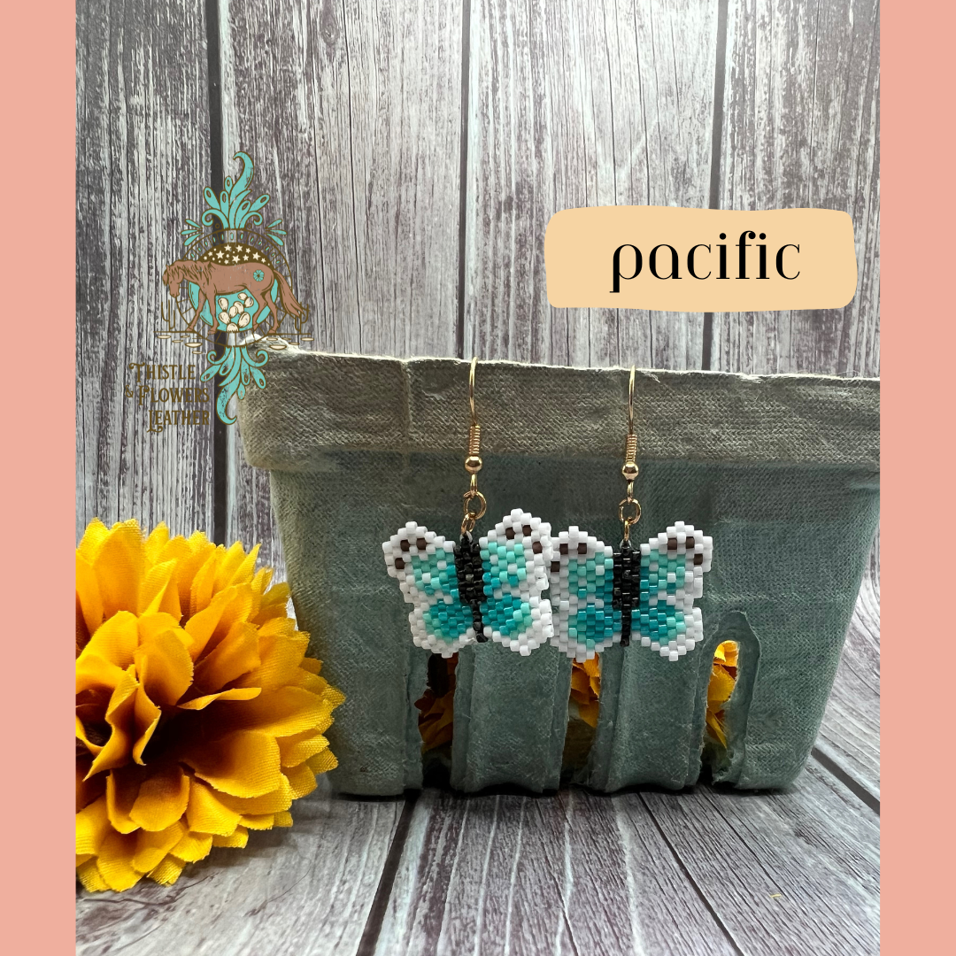 Small beaded earrings in the shape of a butterfly. Bead colors are light turquoise, turquoise, brown, black, and white. Looks like a pixelated butterfly. Gold colored earring hooks and jump rings. Earrings are hanging on a vintage green paper produce container, with a yellow fake marigold flower next to it. Backdrop is gray wood. “Pacific” is in a text box with peach background to identify style, and the Thistle & Flowers Leather logo is on the flower and produce container.