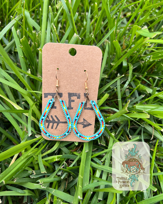Teardrop beaded earrings with turquoise, black, orange, red, yellow, and white beads. Photographed on grass. Logo is in the bottom right corner of photo.