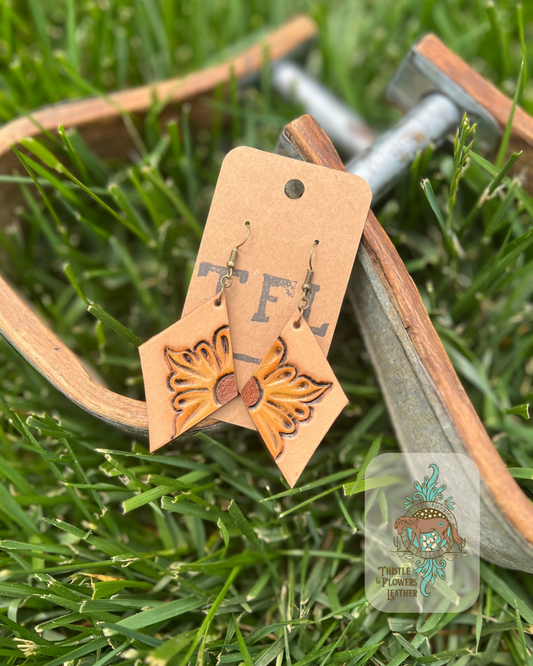 Tooled leather diamond shaped earrings with tooled and painted sunflower yellow flowers on the interior corners of the diamonds. Photographed against metal and wooden stirrups on grass, with logo in bottom right corner of photo.