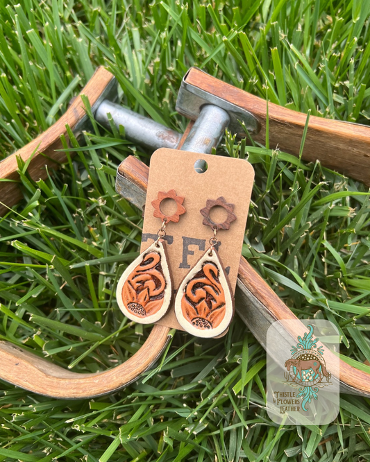 Smaller teardrop earrings hanging from a wooden flower stud (actual stud- in-ear part is not wooden) with a handtooled and painted western design. Features a cream border, sunflower at bottom, and traditional western tooling. Earrings are resting on metal and wooden stirrups on grass. Logo is in bottom right of photo.