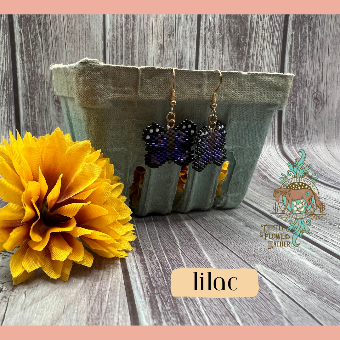 Small beaded earrings in the shape of a butterfly. Bead colors are purple, dark purple, black, brown, and white. Looks like a pixelated butterfly. Gold colored earring hoops and jump rings. Earrings are hanging on a vintage green paper produce container, with a yellow fake marigold flower next to it. Backdrop is gray wood. “Lilac” is in a text box with peach background to identify style, and the Thistle & Flowers Leather logo is on the flower and produce container.