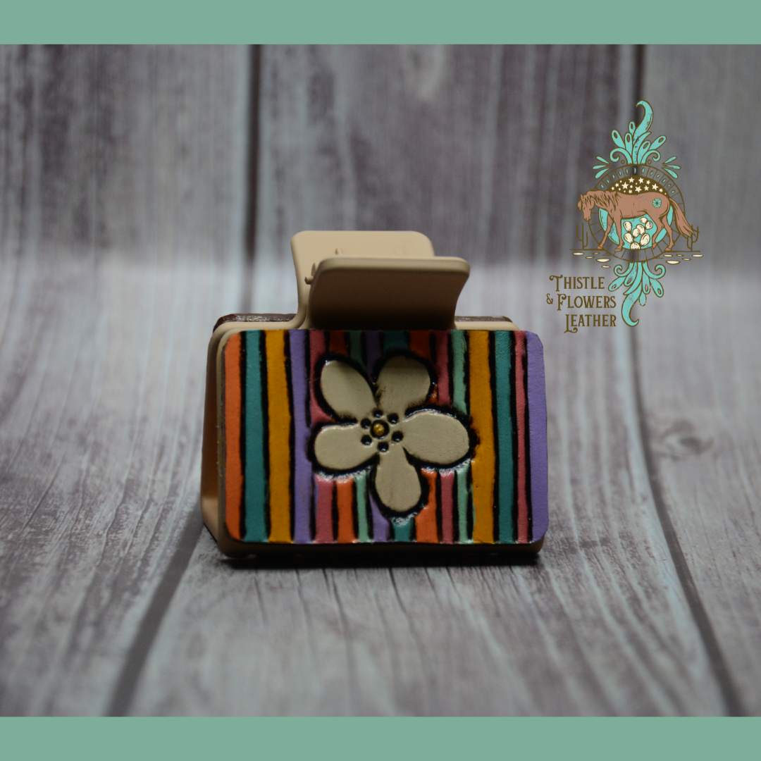 Medium sized plastic hair clip with a tooled and painted patch. Off white flower in the center with colored stripes (orange, turquoise, yellow, purple, pink, and mint)