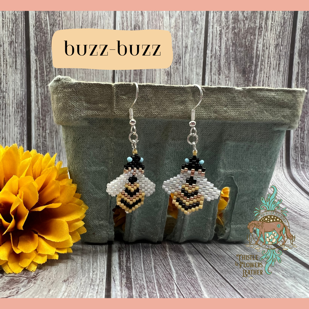 Small beaded earrings in the shape of a bee. Bead colors are yellow, light blue, black, gold, and white. Looks like a pixelated bee. Wings are a slightly see through white. Silver colored earring hooks and jump rings. Earrings are hanging on a vintage green paper produce container, with a yellow fake marigold flower next to it. Backdrop is gray wood. “Buzz buzz” is in a text box with peach background to identify style, and the Thistle & Flowers Leather logo is on the corner of the produce container.
