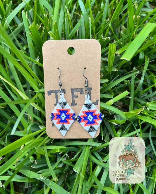 Beaded earrings in a diamond shape with a southwestern design. Bead colors are orange, white, blue, and gunmetal. Photographed on grass with logo in bottom right.