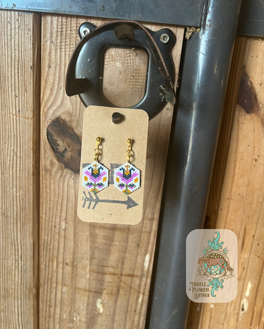 Hand beaded hexagon earrings dangling from a gold colored stud post. Earrings have a vintage tulip design. Bead colors are white, purple, blue, gold, yellow, and green. Earrings are hanging on stall front. Logo is on bottom right corner of photo.