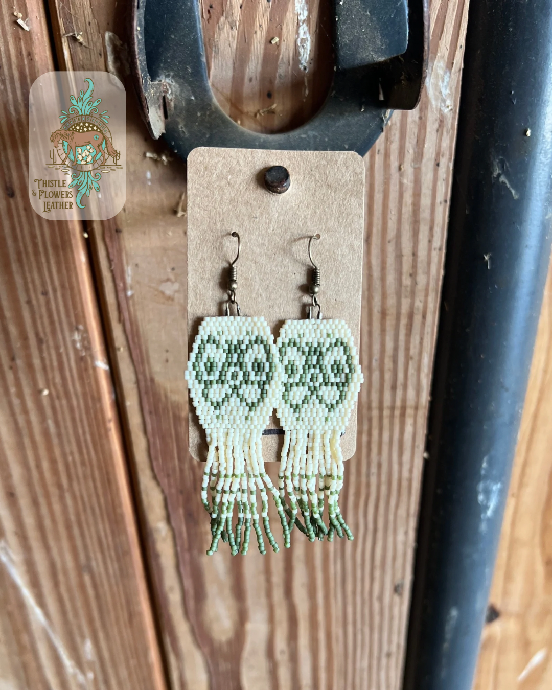 Beaded earrings. Earrings are a hexagon shape with fringe coming off the bottom edge. Beads are cream and sage colored. Sage colored beads create a butterfly design in hexagon shape and add markings on fringe. All beads are matte. Earrings are hanging on stall front. Logo is on the top left corner of photo.