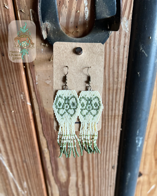Beaded earrings. Earrings are a hexagon shape with fringe coming off the bottom edge. Beads are cream and sage colored. Sage colored beads create a butterfly design in hexagon shape and add markings on fringe. All beads are matte. Earrings are hanging on stall front. Logo is on the top left corner of photo.