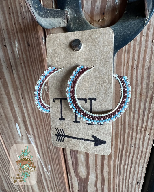 Beaded hoop earrings on silver colored base. Bead colors are copper, turquoise, brown, and cream. Beads make a pattern reminiscent of a western saddle pad. Earrings are hanging on a stall front. Logo is on bottom left corner of photo.