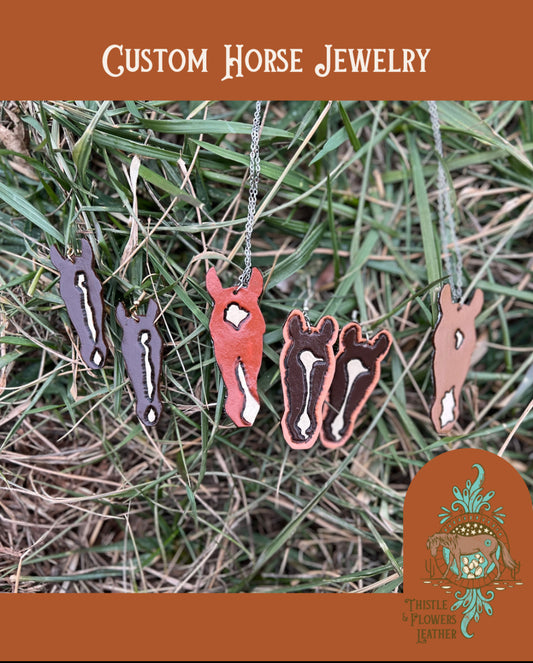 Tooled leather earrings custom tooled and painted to look like individual horses are on clean grass. Custom horse necklaces are also in the photo. Photo has Thistle & Flowers Leather logo watermarked.