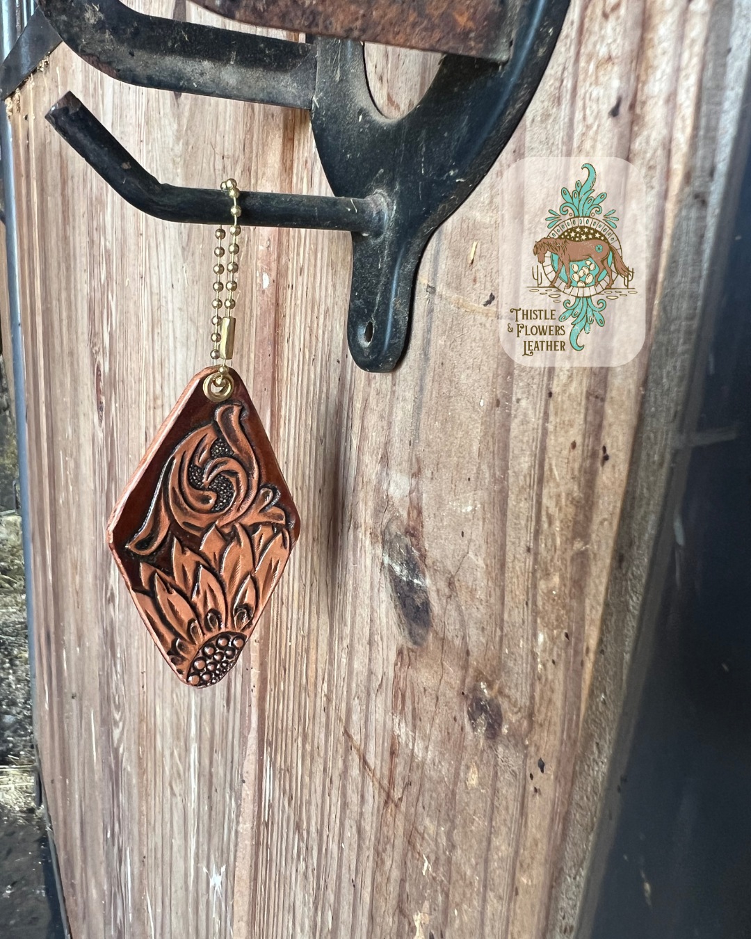 Hotel style keychain, handtooled leather. Features a traditional Western tooling design and a tooled sunflower. Not tooled parts of keychain are dyed brown. Keychain is hanging from stall front. Logo is on the top right of photo. 