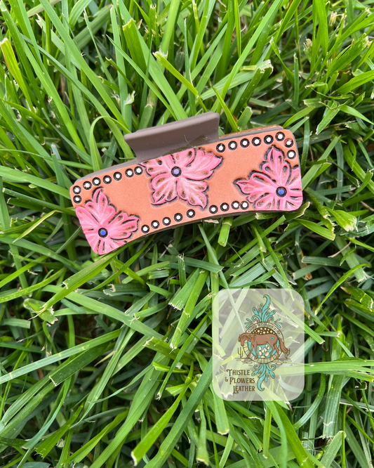 Brown Hairclip base with 3 tooled and painted pink flowers with purple center dots. White dotted tooled and painted border around leather patch. Photographed on grass with logo on bottom right of photo.