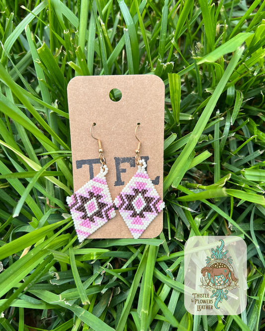 Diamond shaped Hand beaded earrings with cream, various shades of pink, and brown beads in a southwestern design. Photographed on grass with logo on bottom right of photo.