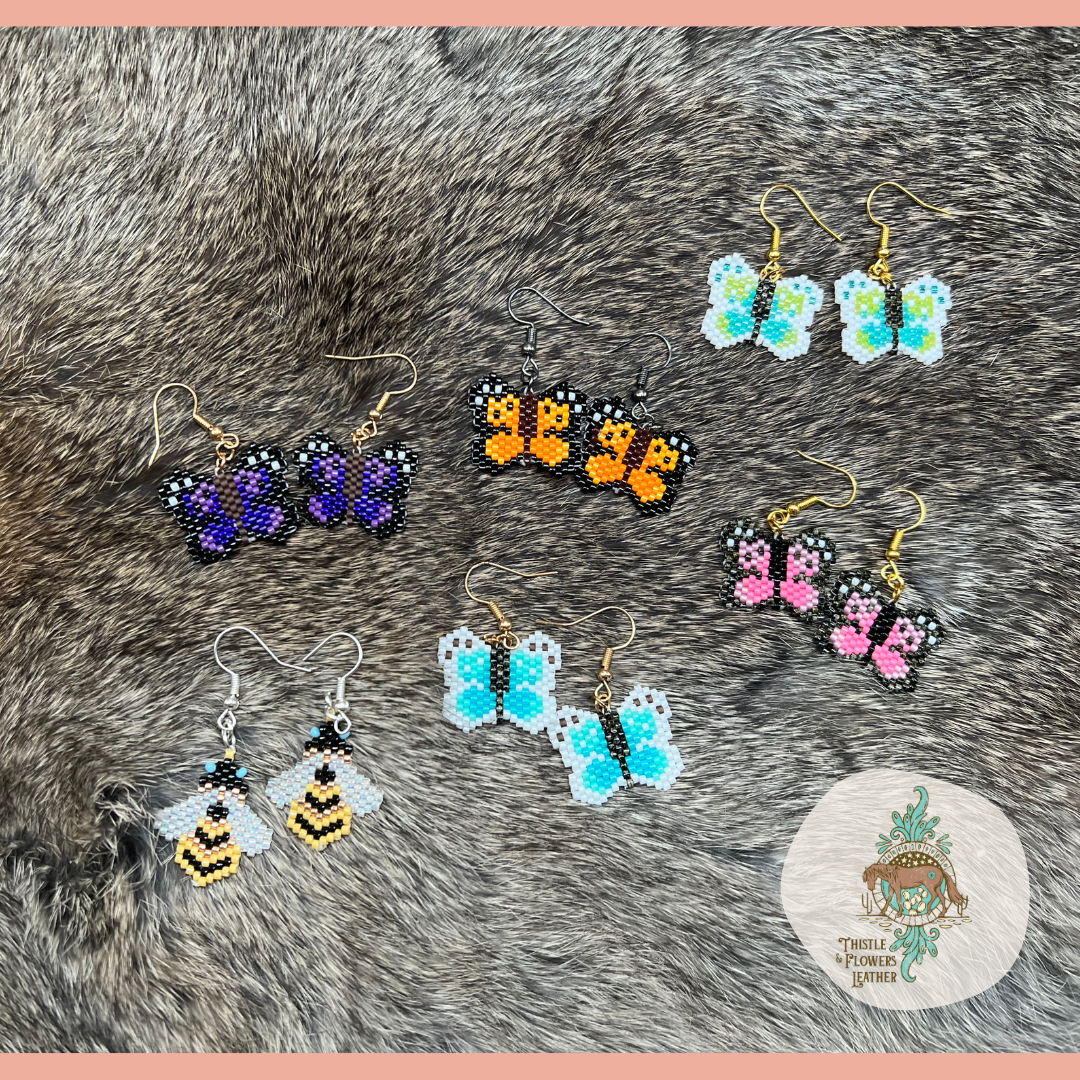 All the beaded butterfly earrings are laying on rabbit fur, with emerald on top, next row from left to right is lilac, monarch, and rose. Next row from left to right is buzz buzz and pacific. Thistle and flowers leather logo is in bottom right corner as a watermark.