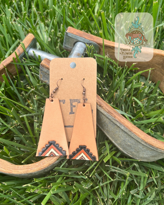 Long trapezoid shaped tooled leather earrings with a western inspired design on the bottom, tooled and painted black, rust, and white. Photographed against metal and wooden stirrups on grass. Logo is in top right corner of photo.