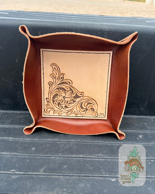 Leather valet tray with a tooled leather base. Base has a hand tooled traditional western floral design. Tray is made up of a reddish brown leather. Base is stitched on with dark thread. Tray is placed on step of mounting block. Logo is in the bottom right of photo.