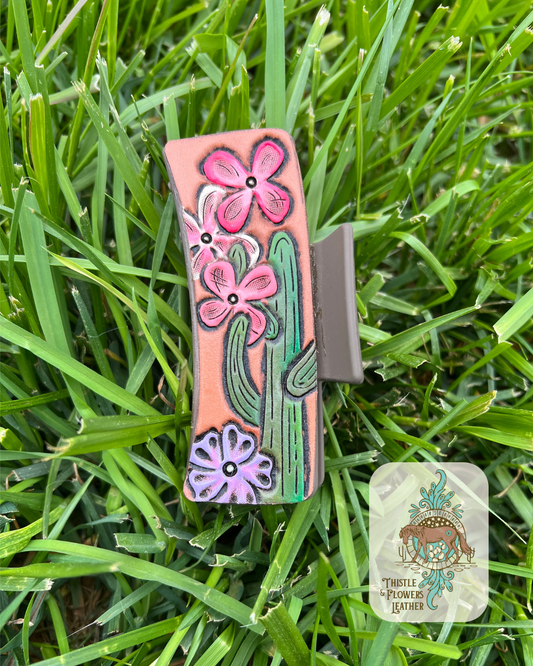 Brown base clip with tooled and painted green cactus, 2 pink flowers, 1 white and pink flower, and 1 purple flower. Photographed on grass with logo on bottom right of photo.