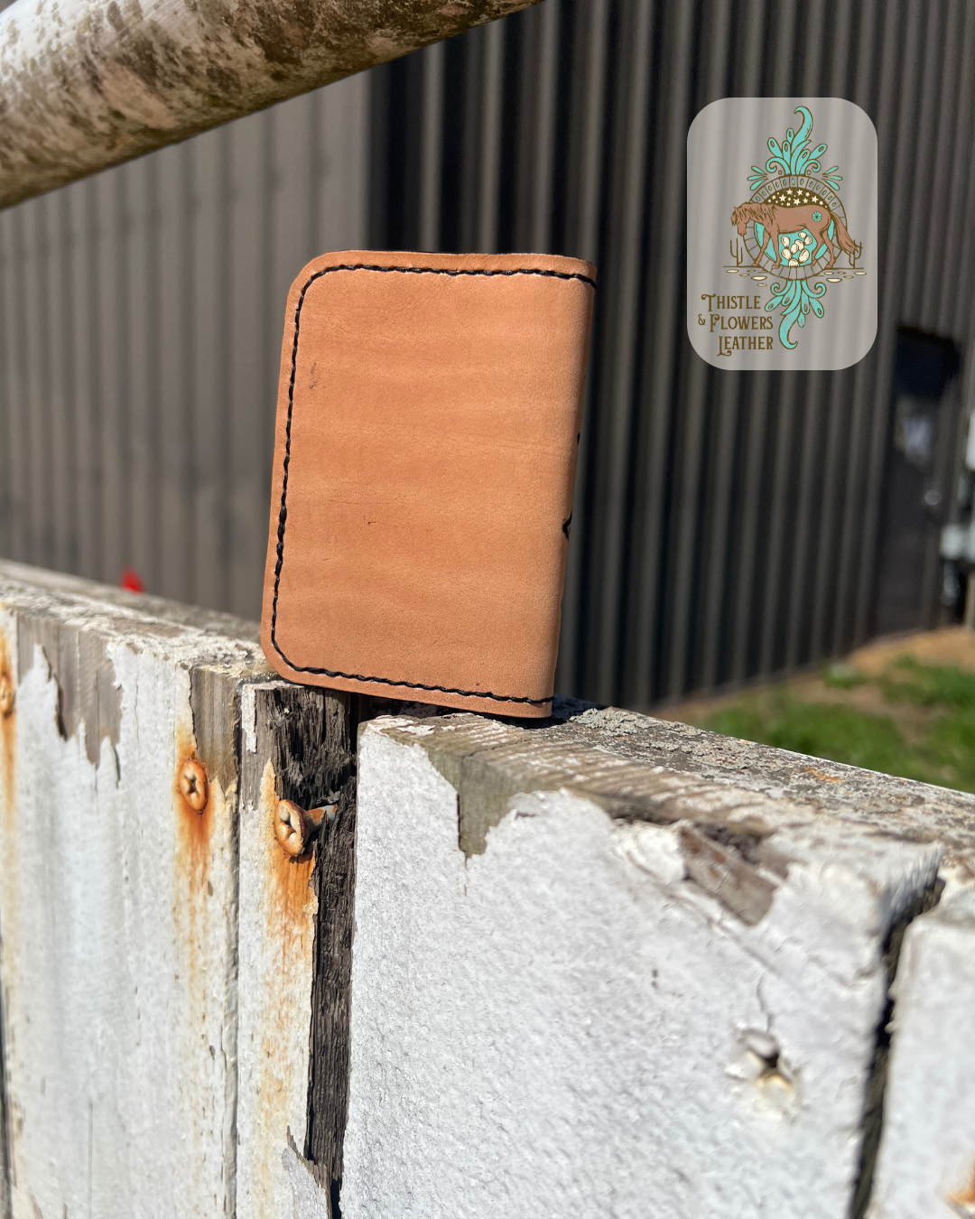 Back of cardholder showing smooth finished leather and stitching. Cardholder is on arena fence. Logo is in top right of photo.