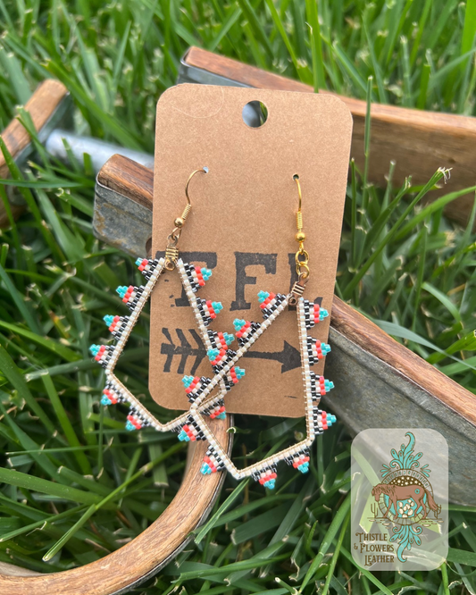 Diamond shaped earrings with beading around edges, with the beadwork creating triangles around the exterior. Bead colors are cream, gunmetal, orange, and turquoise beads. Photographed on metal and wood stirrups with a grass background. Logo is in the bottom right corner of photo.